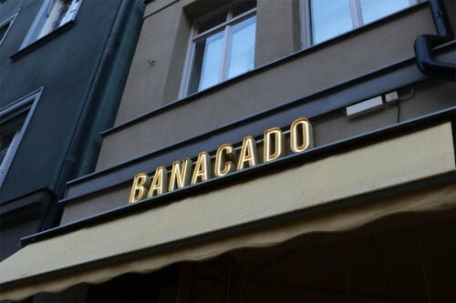 Elegant fasadskylt med texten 'BANACADO' i upplysta bokstäver, monterad ovanför en gul markis och placerad på en klassisk byggnadsfasad.